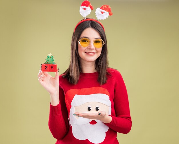 Sonriente joven bastante caucásica vistiendo suéter de santa claus y diadema con gafas sosteniendo y apuntando al juguete del árbol de Navidad con fecha
