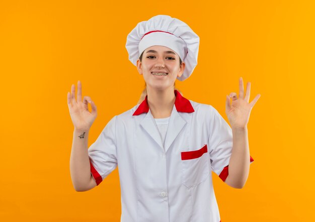 Sonriente joven bastante cocinero en uniforme de chef con aparatos dentales haciendo signos de ok aislados en el espacio naranja