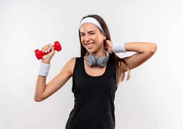 Sonriente joven bastante deportiva con diadema y muñequera y auriculares en el cuello levantando pesas poniendo la mano en el cuello aislado en el espacio en blanco