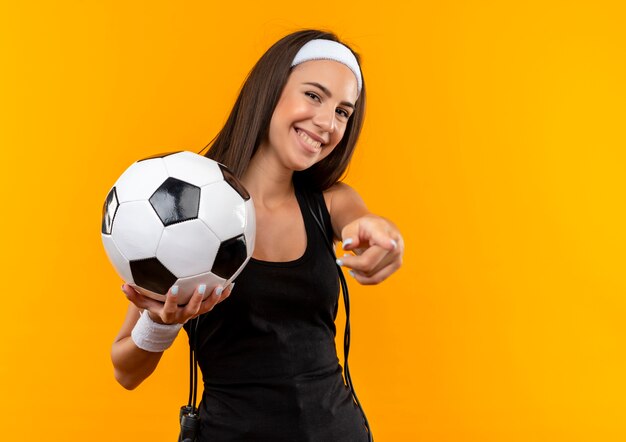 Sonriente joven bastante deportiva con diadema y muñequera sosteniendo un balón de fútbol y apuntando con saltar la cuerda alrededor de su cuello aislado en el espacio naranja