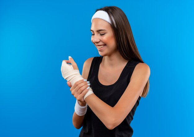 Sonriente joven bastante deportiva con diadema y muñequera sosteniendo y mirando su muñeca lesionada en vendaje aislado en espacio azul