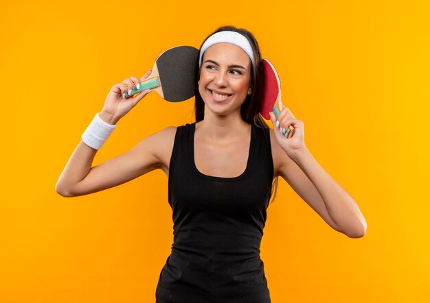 Sonriente joven bastante deportiva con diadema y muñequera tocando su cabeza con raquetas de ping pong y mirando al lado aislado en el espacio naranja