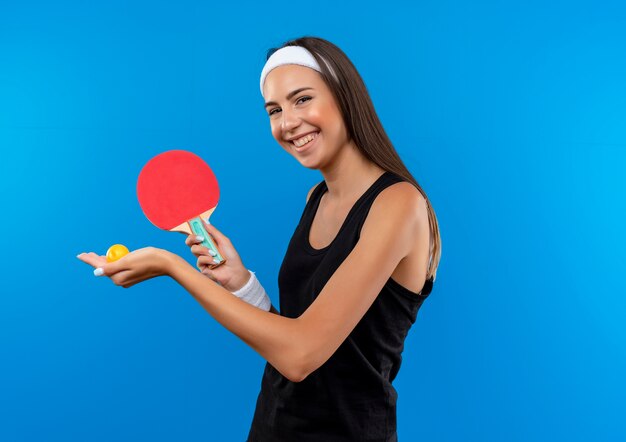 Sonriente joven bastante deportiva vistiendo diadema y muñequera sosteniendo raquetas de ping pong y pelota en el espacio azul