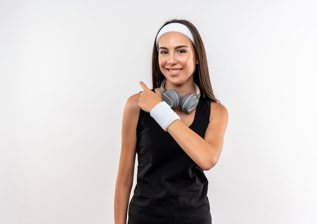 Sonriente joven bastante deportivo vistiendo diadema y muñequera y auriculares en el cuello apuntando hacia atrás en el espacio en blanco