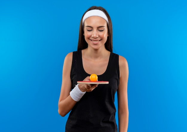 Sonriente joven bastante deportivo vistiendo diadema y muñequera sosteniendo y mirando la raqueta de ping pong con pelota en el espacio azul