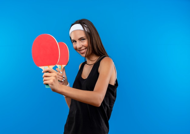Sonriente joven bastante deportivo vistiendo diadema y muñequera sosteniendo raquetas de ping pong aisladas en el espacio azul