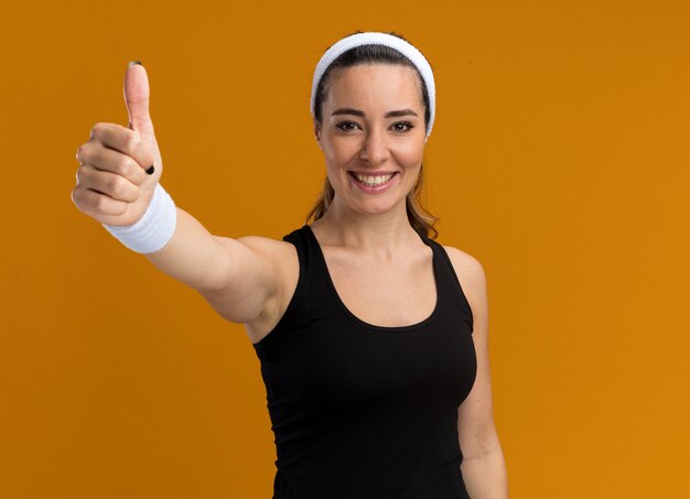 Sonriente joven bastante deportivo vistiendo diadema y muñequeras mostrando el pulgar hacia arriba