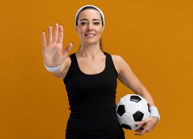 Sonriente joven bastante deportivo vistiendo diadema y muñequeras sosteniendo un balón de fútbol haciendo gesto de parada