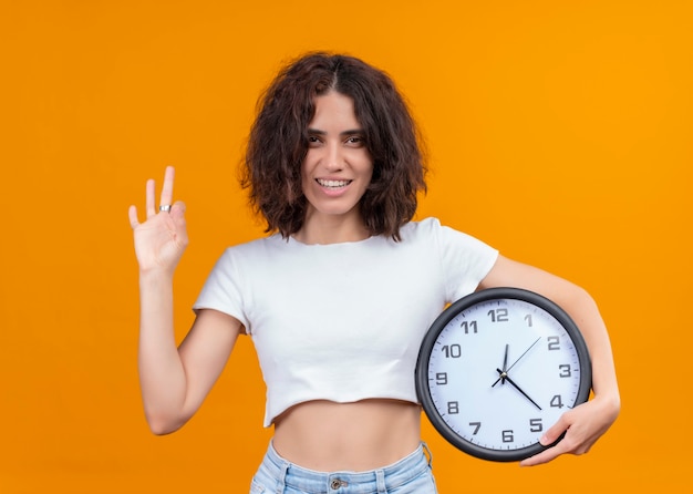 Sonriente joven bella mujer sosteniendo el reloj y haciendo bien firmar en la pared naranja aislada