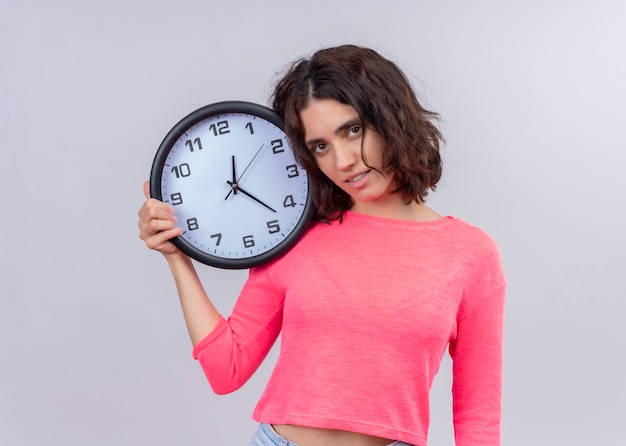 Sonriente joven bella mujer sosteniendo el reloj en la pared blanca aislada con espacio de copia