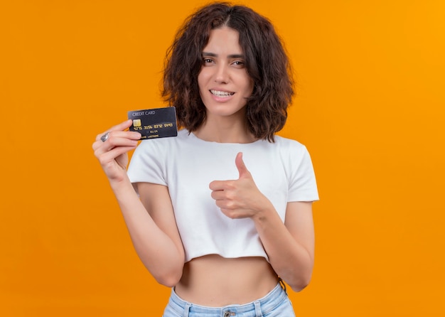 Sonriente joven bella mujer sosteniendo la tarjeta y mostrando el pulgar hacia arriba en la pared naranja aislada con espacio de copia