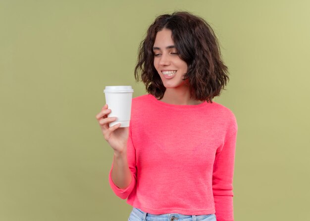 Sonriente joven bella mujer sosteniendo una taza de café de plástico y mirándolo en la pared verde aislada con espacio de copia