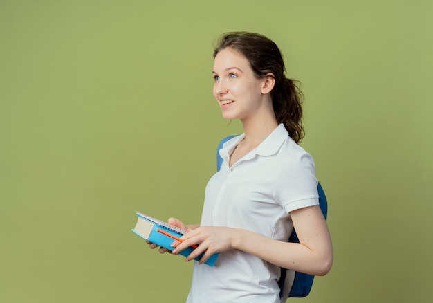 Sonriente joven bonita estudiante vistiendo bolsa trasera de pie en la vista de perfil sosteniendo el bloc de notas y el libro mirando directamente