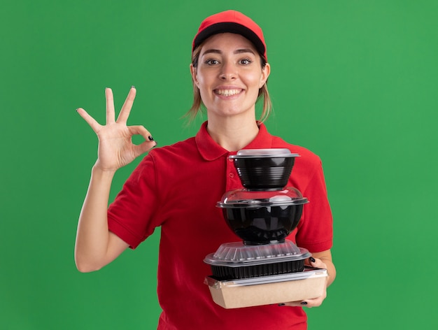 Sonriente joven bonita repartidora en gestos uniformes signo de mano ok y sostiene contenedores de comida en paquete de comida en verde