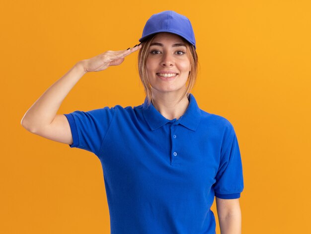 Sonriente joven bonita repartidora en uniforme haciendo gesto de saludo en naranja