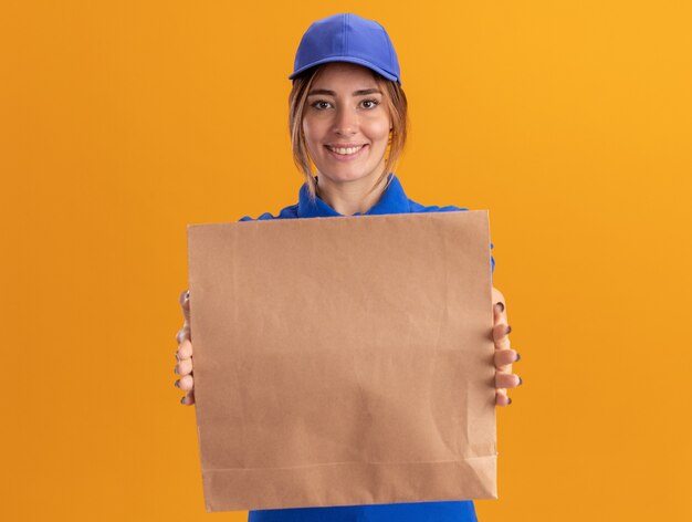 Sonriente joven bonita repartidora en uniforme con paquete de comida de papel en naranja