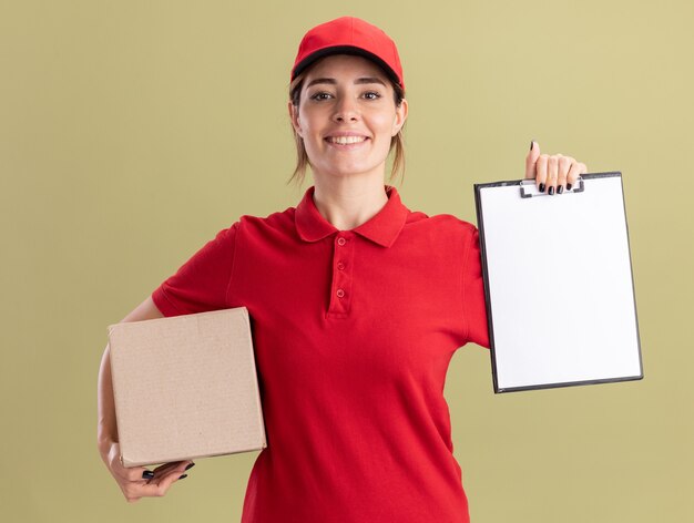 Sonriente joven bonita repartidora en uniforme con portapapeles y caja de cartón en verde oliva