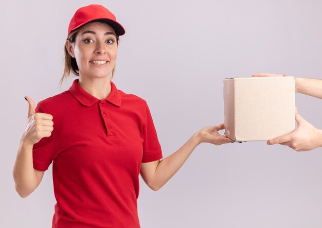 Sonriente joven bonita repartidora en uniforme pulgar hacia arriba y da caja de cartón a alguien en blanco