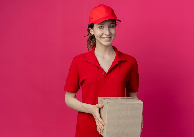 Sonriente joven bonita repartidora en uniforme rojo y gorra con caja de cartón aislada sobre fondo carmesí con espacio de copia