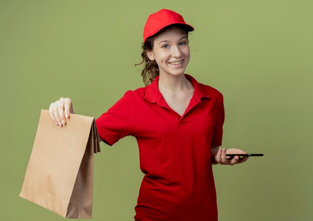 Sonriente joven bonita repartidora en uniforme rojo y gorra estirando el paquete de papel aislado sobre fondo verde oliva