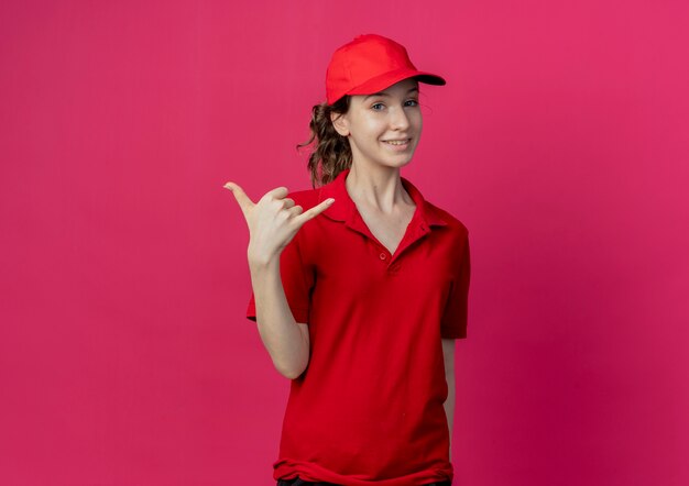 Sonriente joven bonita repartidora en uniforme rojo y gorra haciendo gesto suelto aislado sobre fondo carmesí con espacio de copia