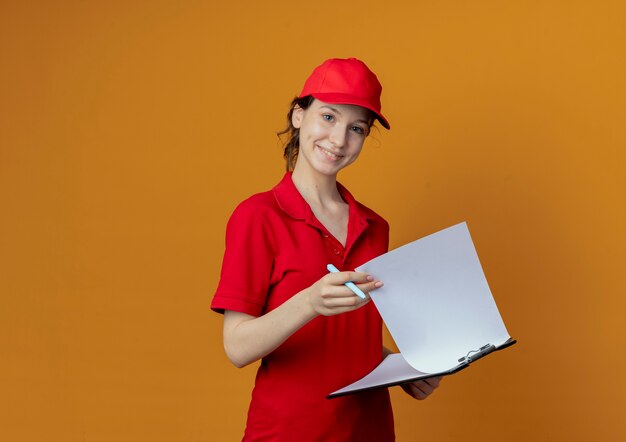 Sonriente joven bonita repartidora en uniforme rojo y gorra sosteniendo portapapeles y bolígrafo aislado sobre fondo naranja con espacio de copia