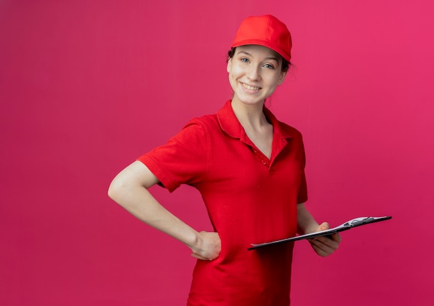 Sonriente joven bonita repartidora en uniforme rojo y gorra sosteniendo el portapapeles poniendo la mano en la cintura aislada sobre fondo carmesí con espacio de copia