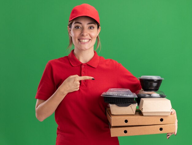 Sonriente joven bonita repartidora en uniforme sostiene y apunta a paquetes de alimentos de papel y contenedores en cajas de pizza en verde