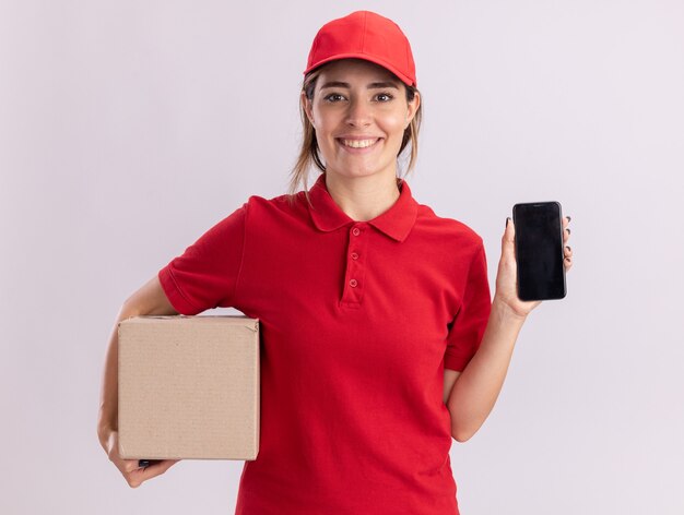 Sonriente joven bonita repartidora en uniforme sostiene cardbox y teléfono en blanco