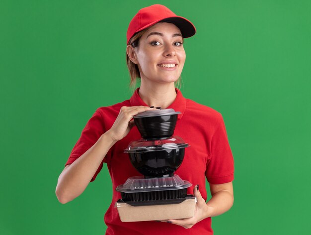 Sonriente joven bonita repartidora en uniforme sostiene contenedores de comida y paquete de comida en verde