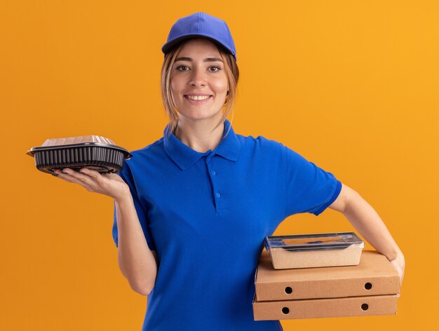 Sonriente joven bonita repartidora en uniforme sostiene paquetes de alimentos de papel y contenedores en cajas de pizza en naranja