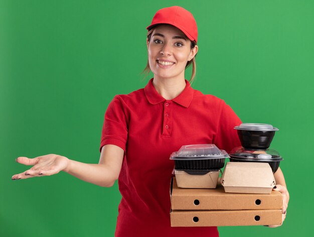 Sonriente joven bonita repartidora en uniforme sostiene tiene paquetes de alimentos de papel y contenedores en cajas de pizza