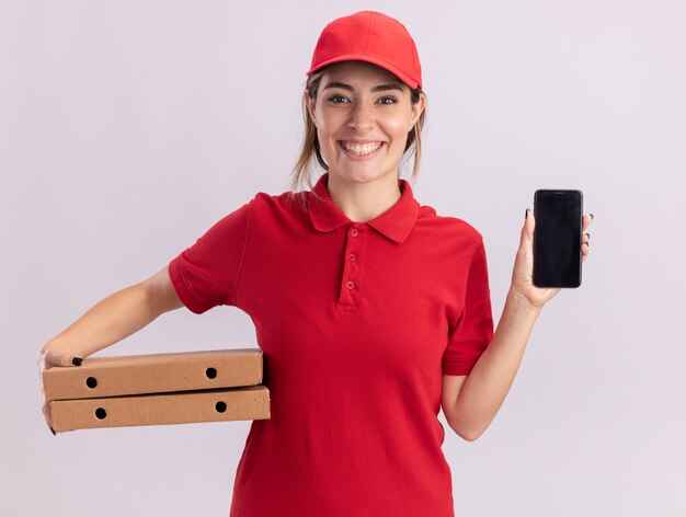 Sonriente joven bonita repartidora en uniforme tiene cajas de pizza y teléfono en blanco