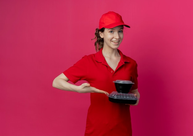 Sonriente joven bonita repartidora vestida con uniforme rojo y gorra sosteniendo y apuntando con la mano a los contenedores de comida aislados sobre fondo carmesí con espacio de copia