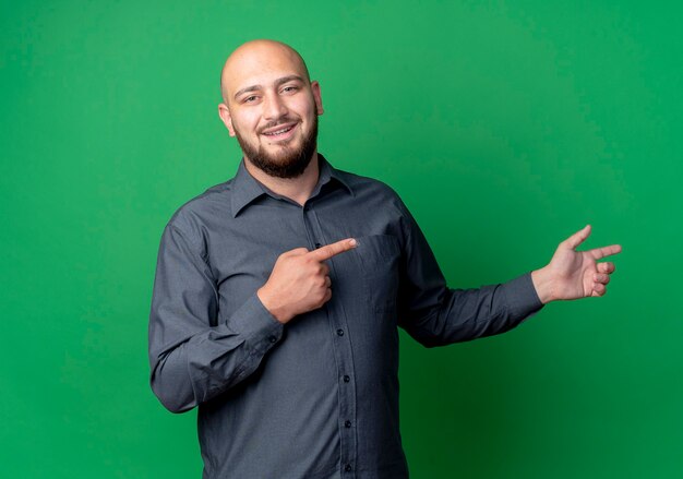 Sonriente joven calvo call center hombre apuntando al lado aislado en la pared verde