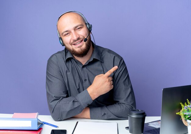 Sonriente joven calvo call center hombre con auriculares sentado en un escritorio con herramientas de trabajo apuntando al lado aislado en la pared púrpura