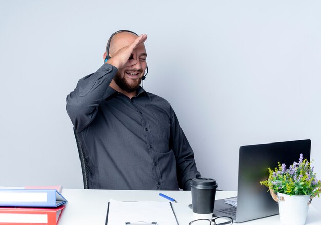 Sonriente joven calvo call center hombre con auriculares sentado en el escritorio con herramientas de trabajo haciendo gesto de mirada en el portátil aislado en la pared blanca