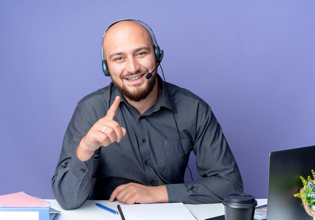 Foto gratuita sonriente joven calvo call center hombre con auriculares sentado en el escritorio con herramientas de trabajo levantando el dedo y poniendo la mano en el escritorio aislado en la pared púrpura