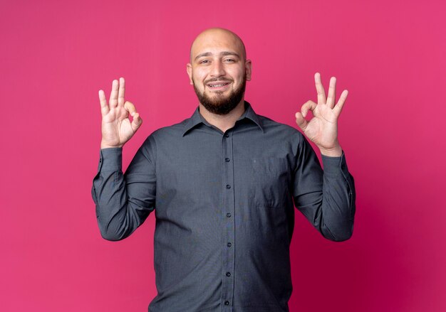 Sonriente joven calvo call center hombre haciendo bien signos aislados en la pared carmesí