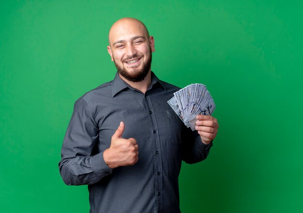 Sonriente joven calvo call center hombre sosteniendo dinero y mostrando el pulgar hacia arriba aislado en la pared verde