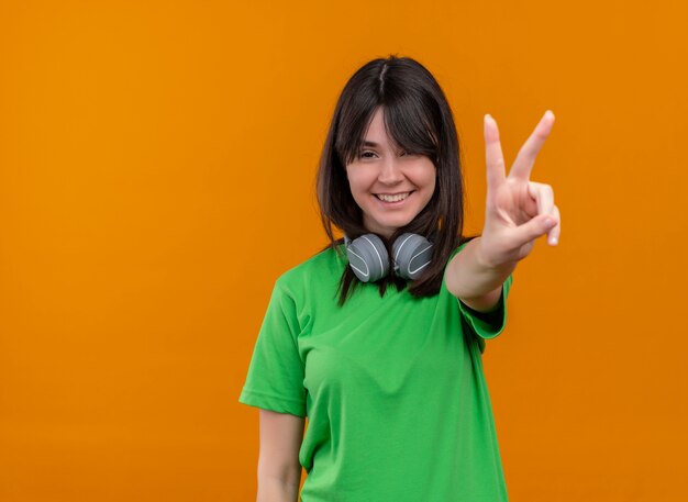 Sonriente joven caucásica en camisa verde con auriculares muestra gesto de la mano de la victoria sobre fondo naranja aislado con espacio de copia