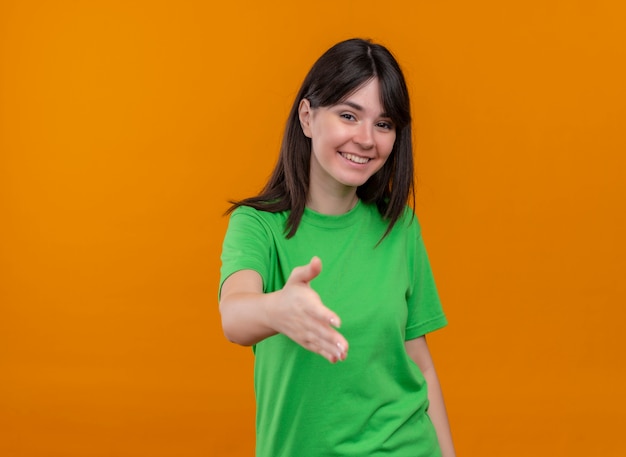 Sonriente joven caucásica en camisa verde extiende la mano y mira a la cámara sobre fondo naranja aislado con espacio de copia