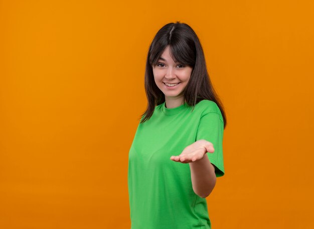 Sonriente joven caucásica en camisa verde muestra la mano vacía a la cámara sobre fondo naranja aislado con espacio de copia