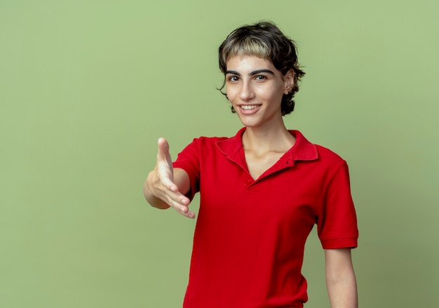 Foto gratuita sonriente joven caucásica con corte de pelo pixie estirando la mano gesticulando hola aislado sobre fondo verde oliva con espacio de copia