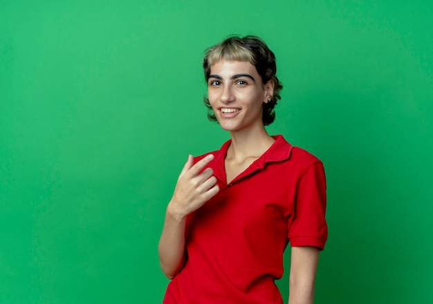 Sonriente joven caucásica con corte de pelo pixie manteniendo la mano en el aire aislado sobre fondo verde con espacio de copia