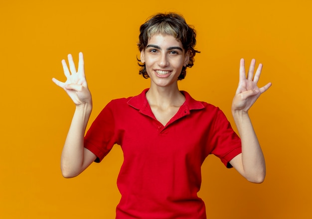 Foto gratuita sonriente joven caucásica con corte de pelo pixie mostrando ocho con manos aisladas sobre fondo naranja