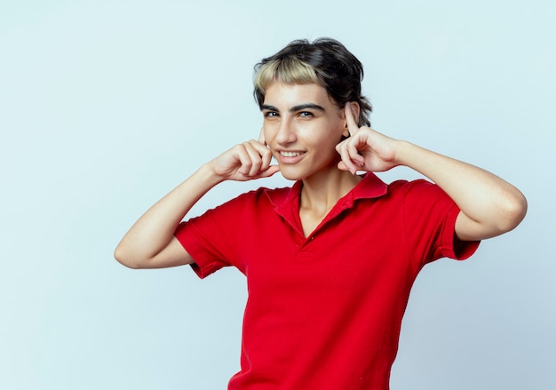 Sonriente joven caucásica con corte de pelo pixie poniendo los dedos en las orejas aislado sobre fondo blanco.