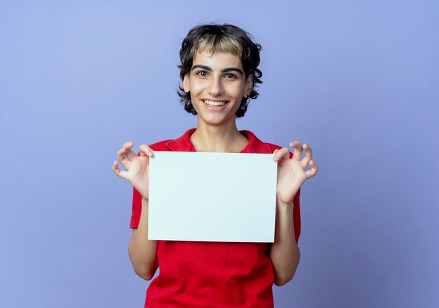 Sonriente joven caucásica con corte de pelo pixie sosteniendo papel aislado sobre fondo púrpura con espacio de copia