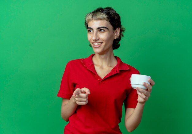 Sonriente joven caucásica con corte de pelo pixie sosteniendo la taza mirando a un lado y apuntando a la cámara aislada sobre fondo verde con espacio de copia