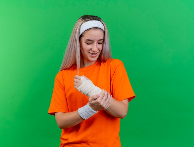 Sonriente joven caucásica deportiva con tirantes con diadema y muñequeras pone la mano y mira el brazo